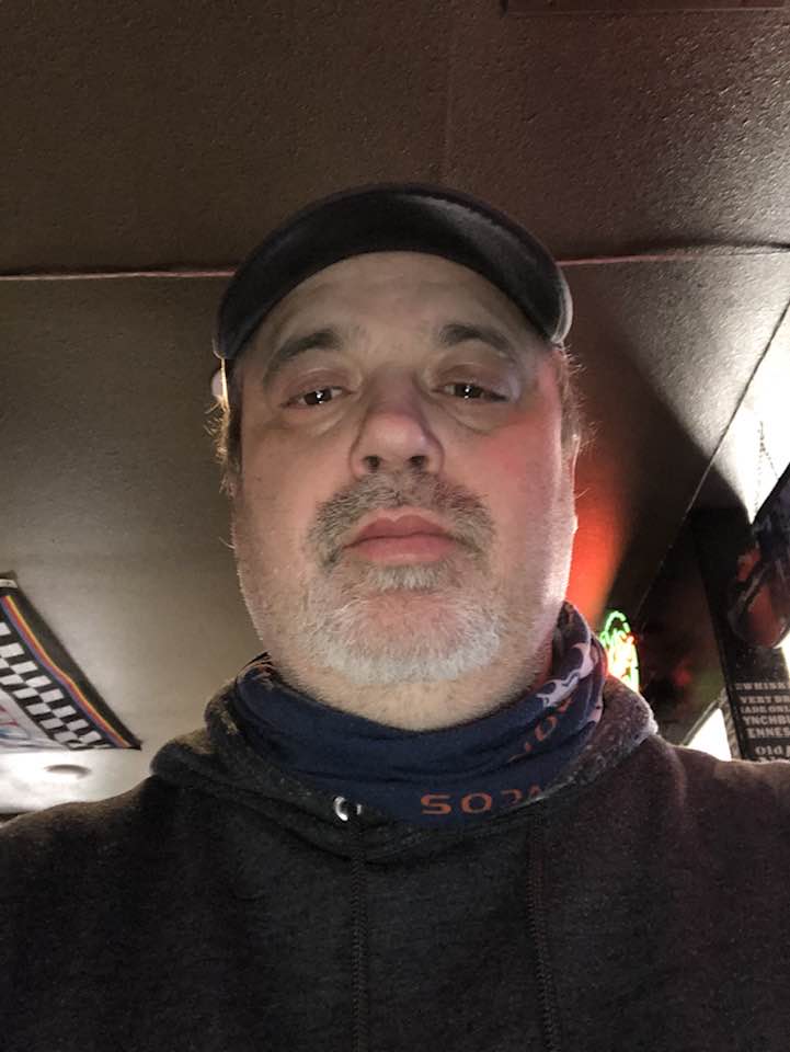 A man wearing a cap and a neck gaiter is taking a selfie indoors with dim lighting.